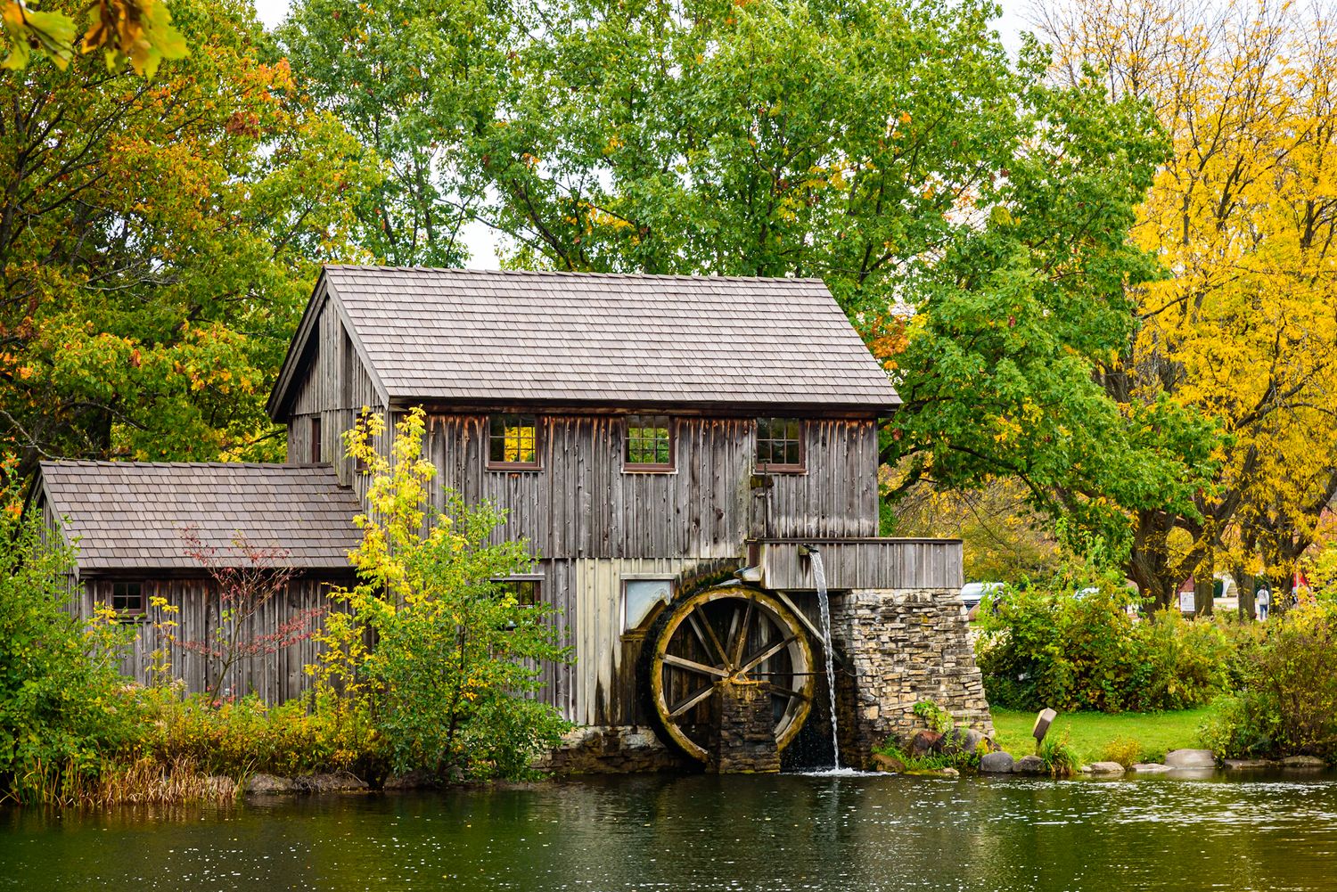 Rustic wooden grist mill with water wheel beside a tranquil pond surrounded by autumn foliage trees.