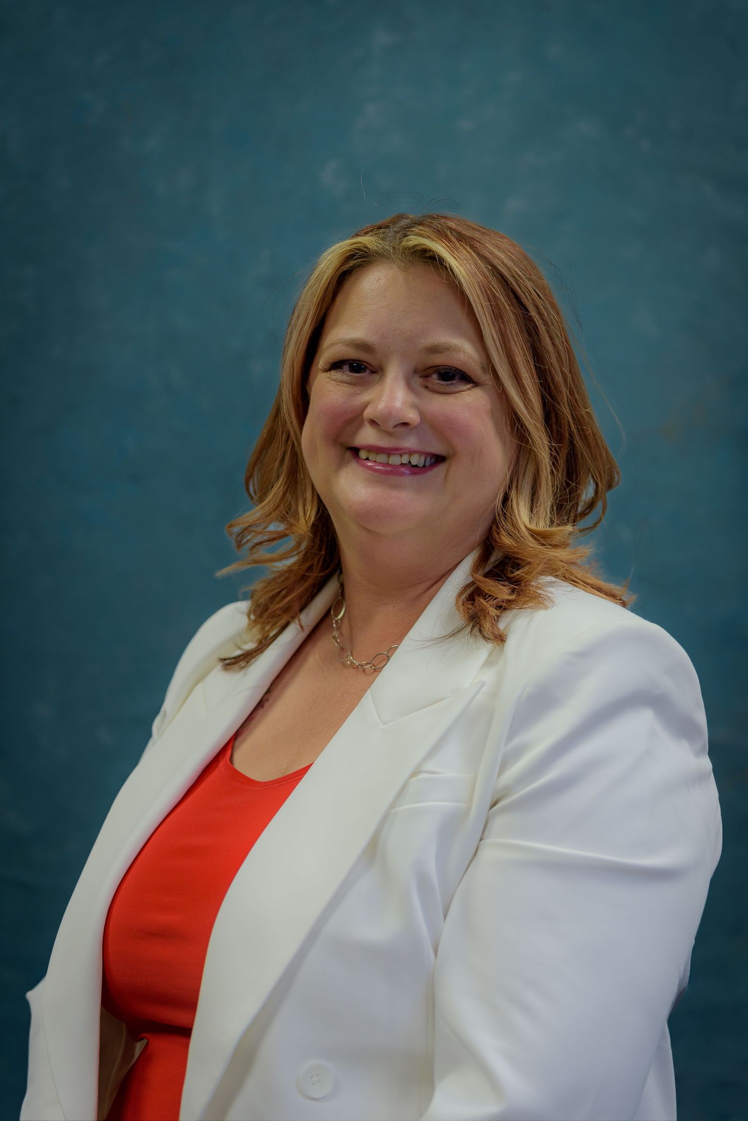 Professional woman in white blazer and red top smiling confidently against a teal background.