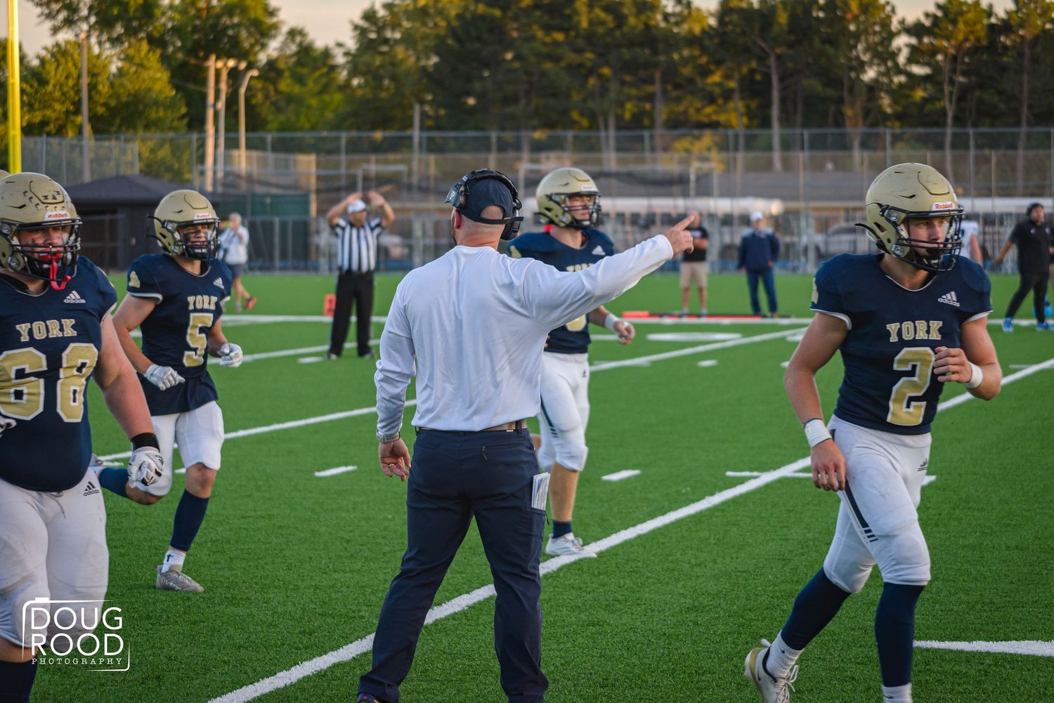 Football coach directing players on field during York high school game, with referee and players in navy uniforms visible.