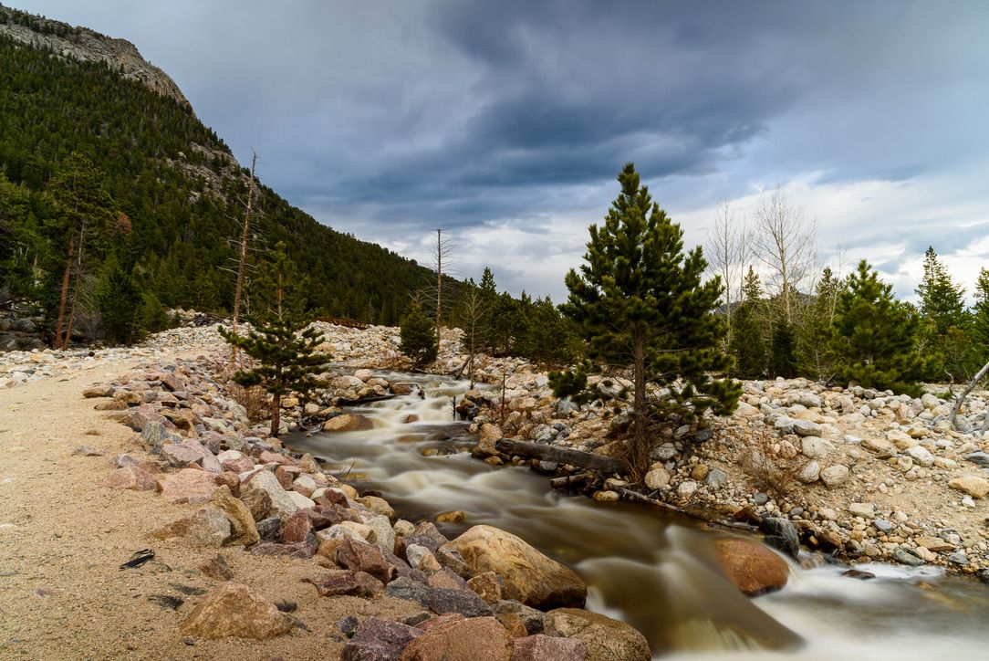 Rocky mountain stream flows through boulder-lined banks with pine trees and dramatic storm clouds in a wilderness landscape.