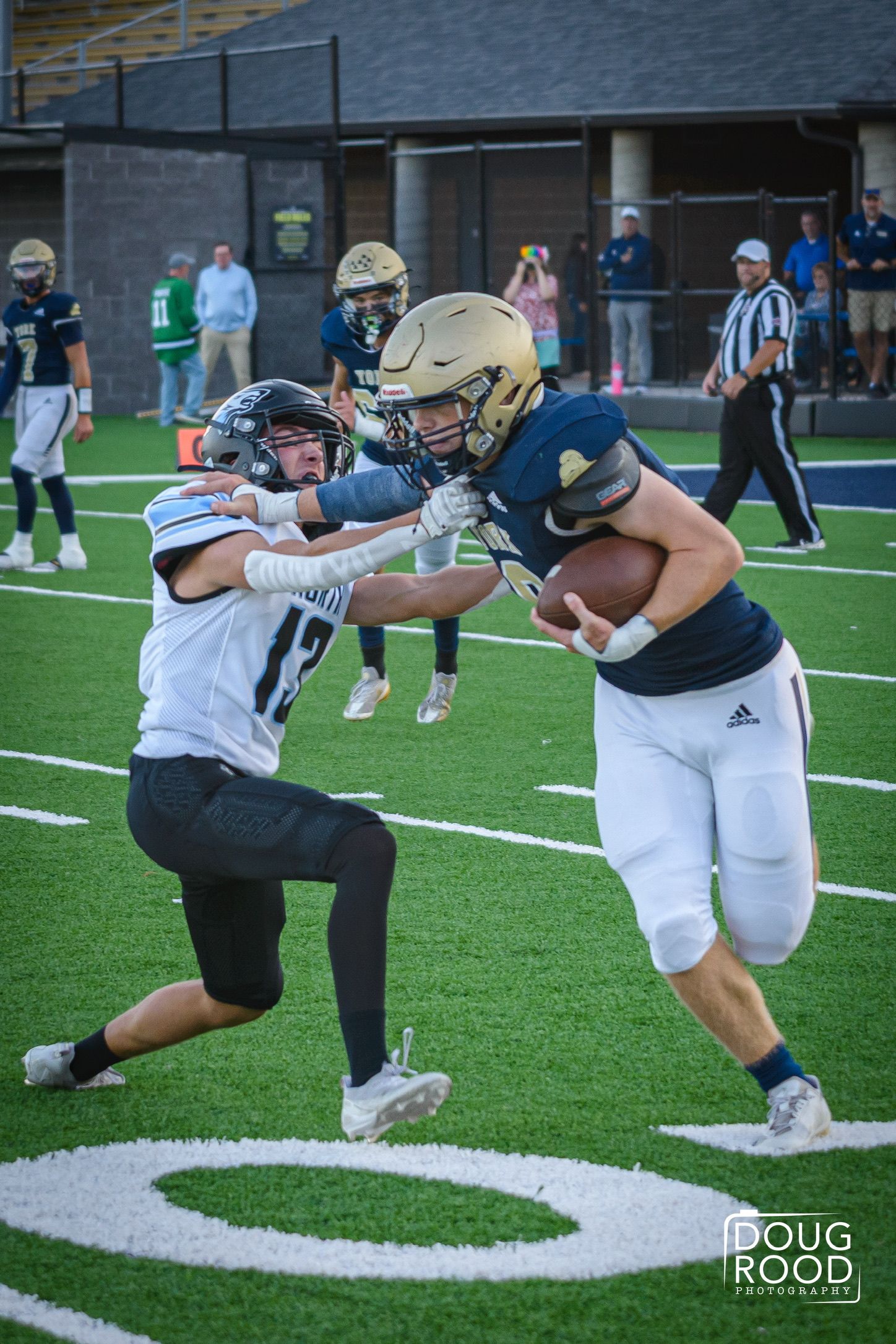 High school football player in navy uniform stiff-arms defender in white jersey during competitive game on turf field.