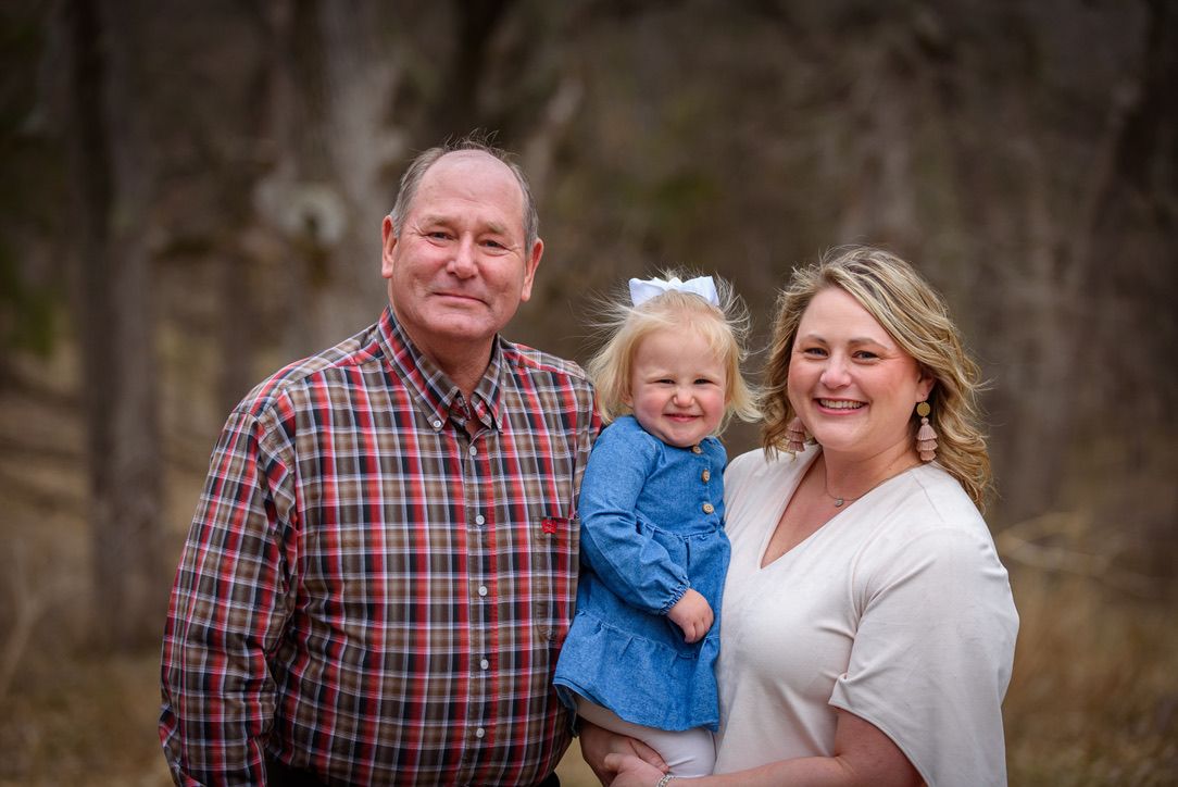 A smiling family of three poses outdoors, with parents holding a young toddler girl in a blue dress among autumn trees.