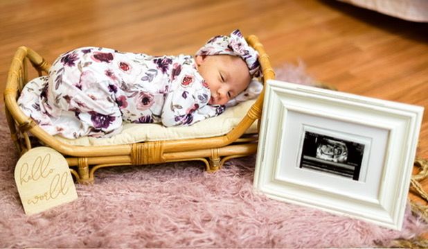 Newborn baby sleeping in floral swaddle in rattan bassinet beside framed ultrasound photo on pink fur rug.