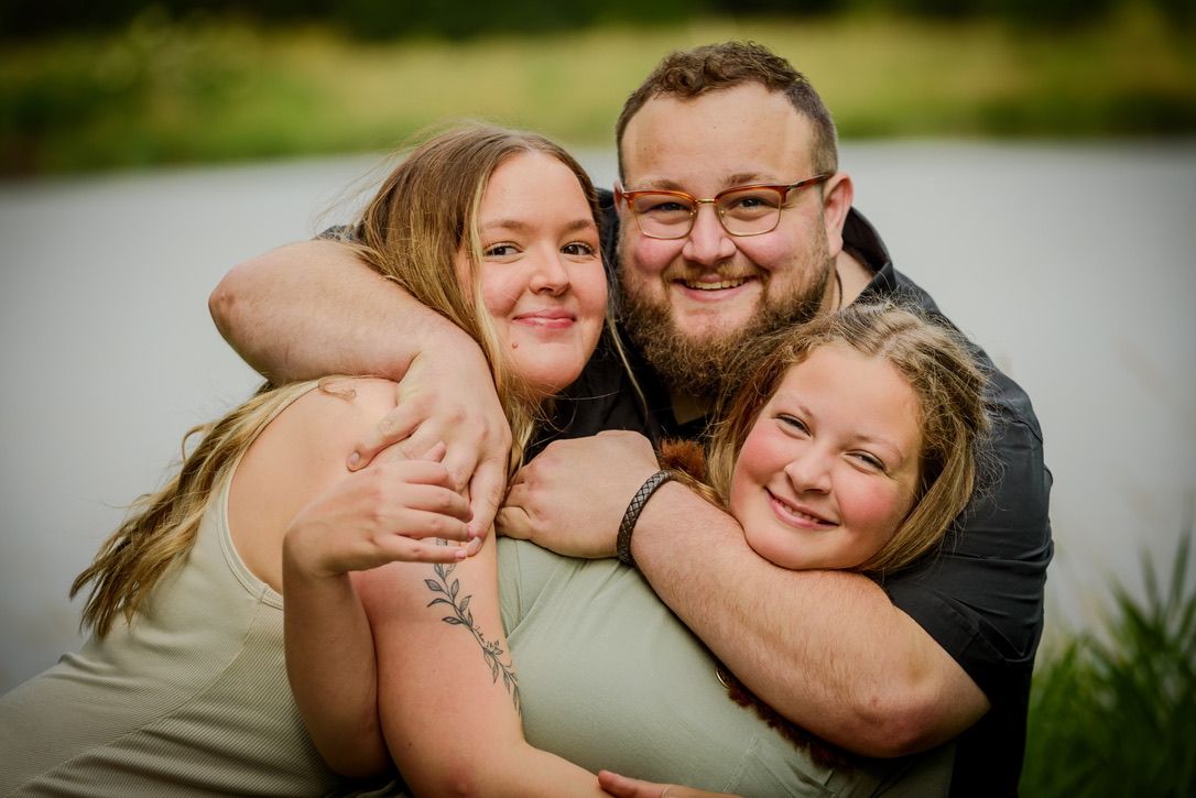 A smiling family of three sharing a warm hug outdoors near a pond, with lush green trees in the background.