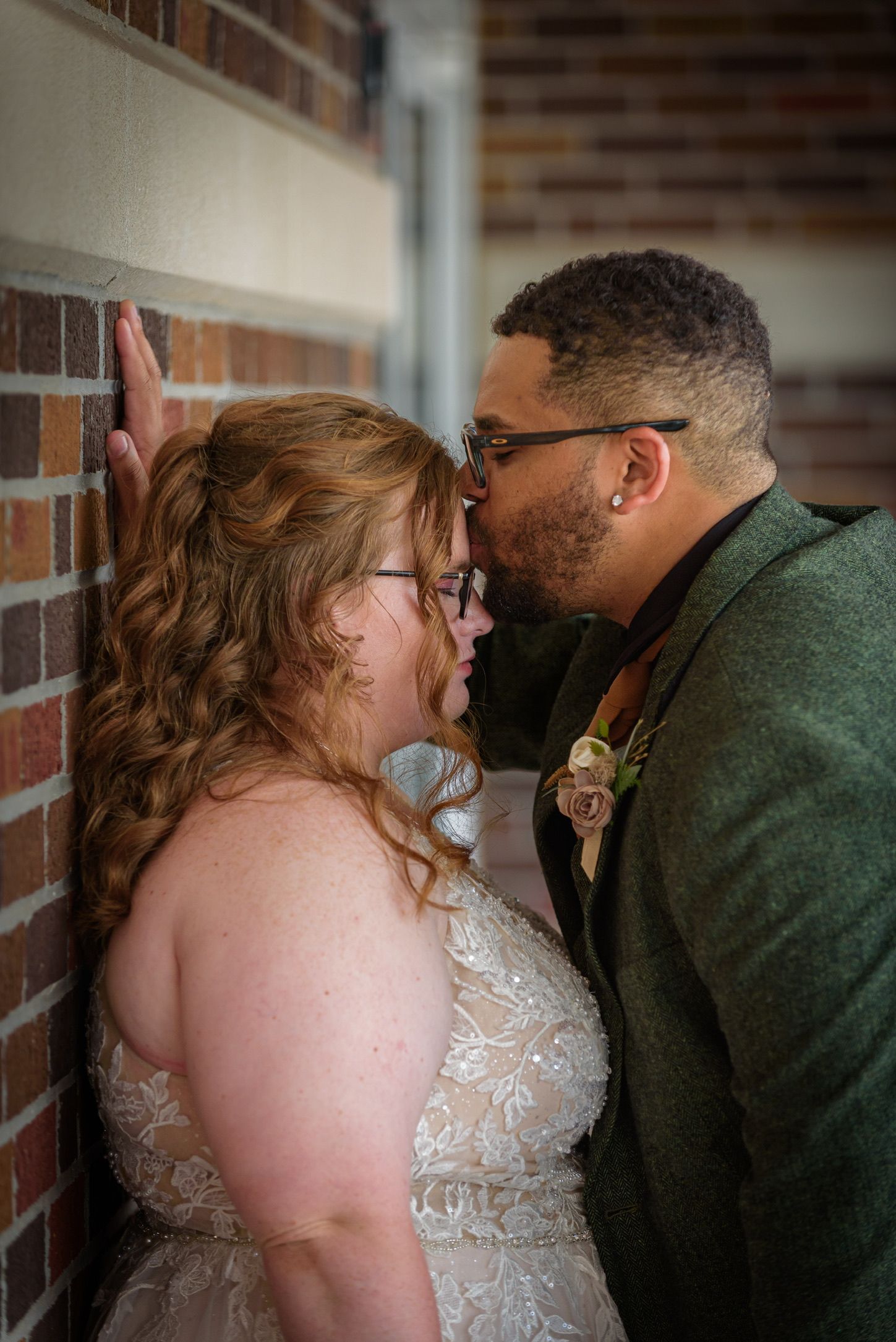 Bride and groom share an intimate moment against a brick wall, she in lace gown, he in green suit with boutonniere.