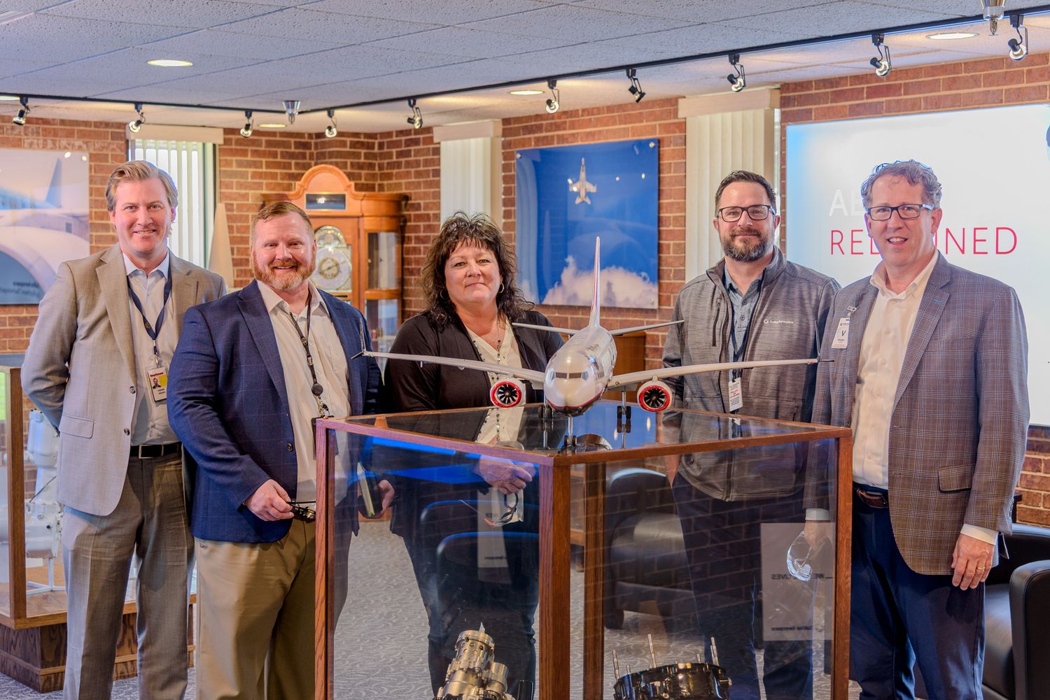 Five professionals pose behind a model airplane display in a museum exhibit with brick walls and framed artwork.