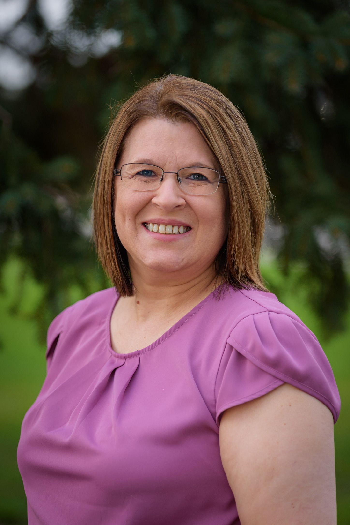 Smiling middle-aged woman with glasses and brown hair wearing a pink blouse, posing outdoors with green trees behind her.
