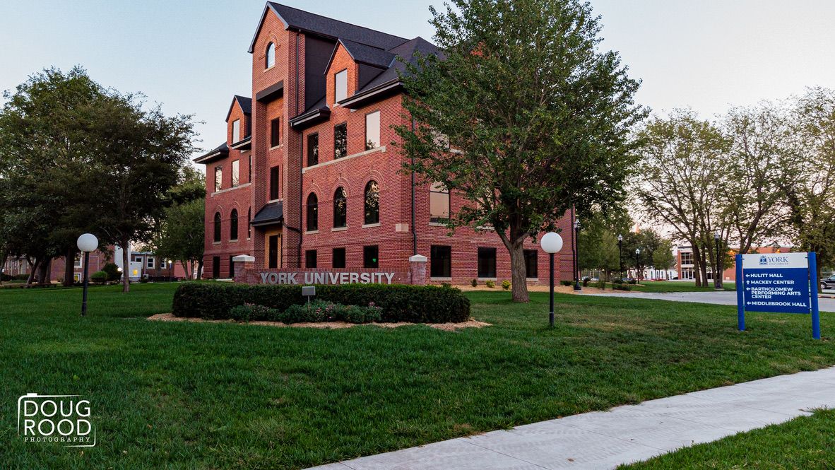 Brick university building with green lawn, trees, and signage reading University on a sunny day.