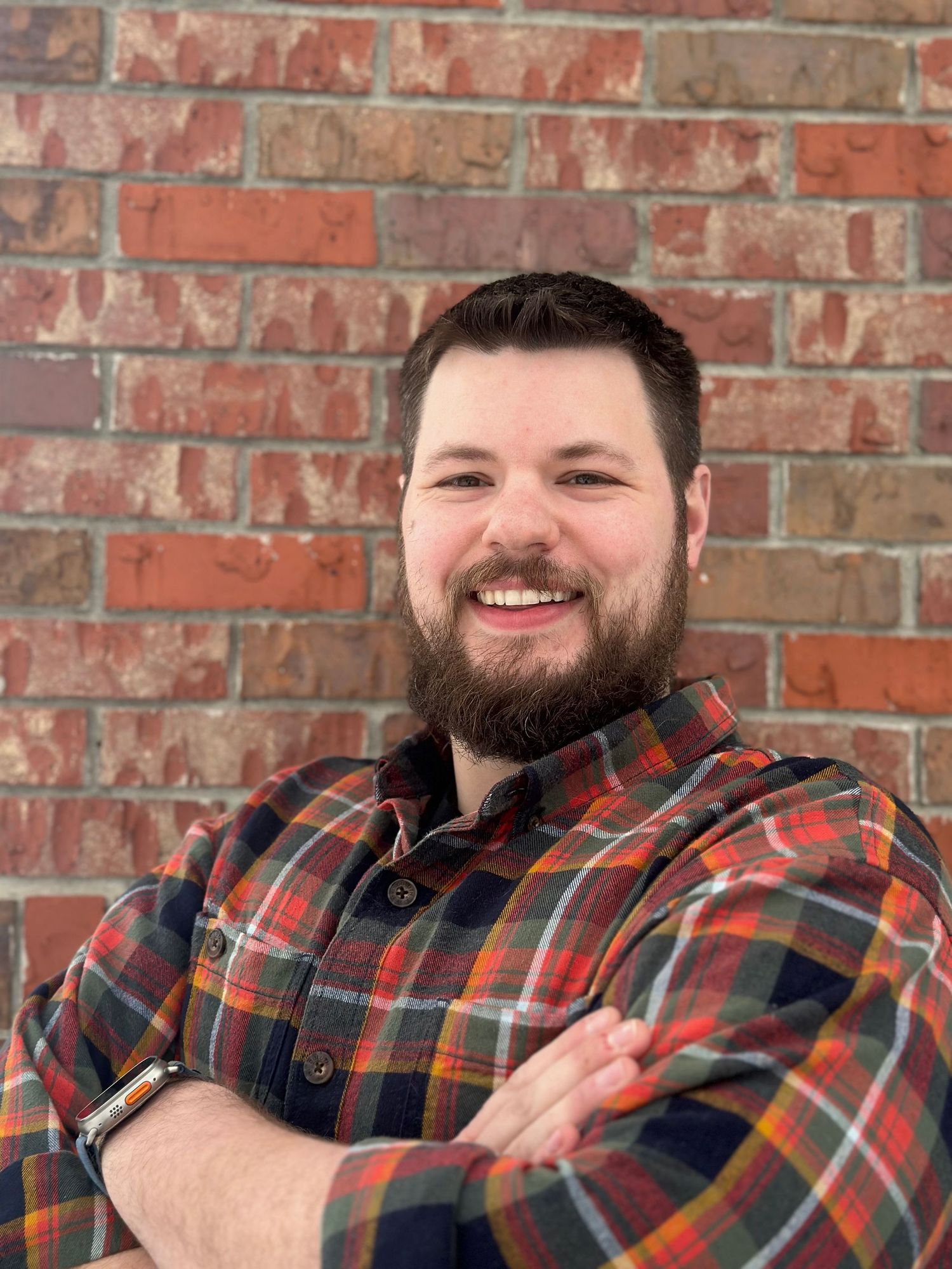 Bearded man smiling with arms crossed against a brick wall wearing a plaid shirt. York, NE