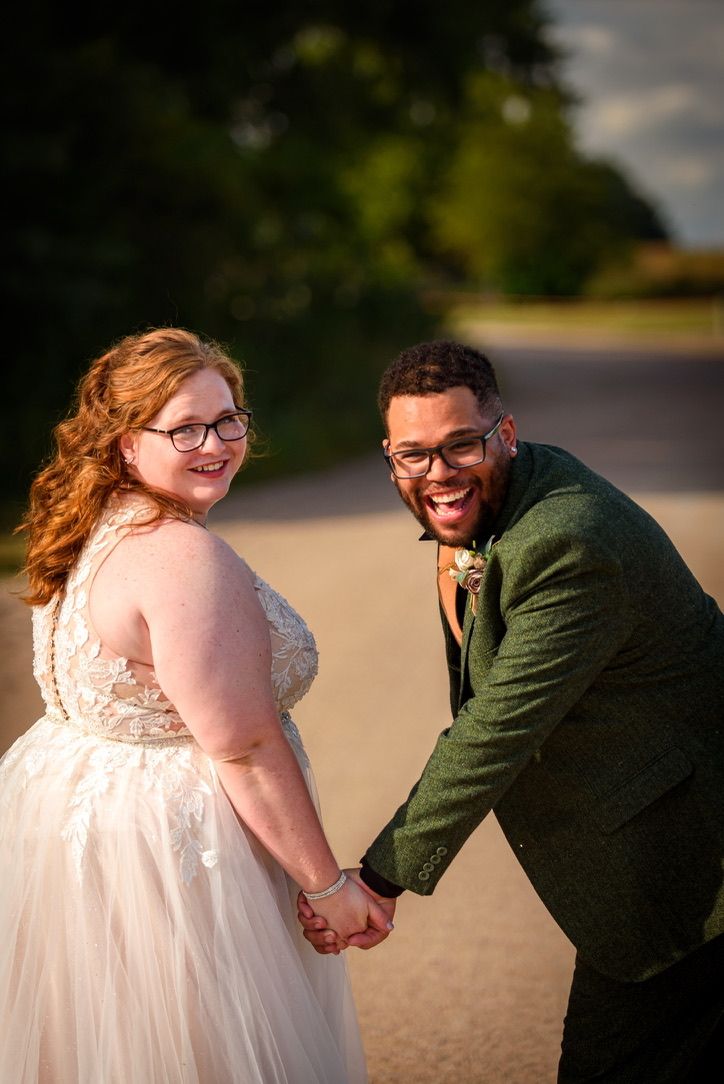 Smiling newlywed couple holds hands on outdoor path, groom in green jacket with boutonniere at golden hour.