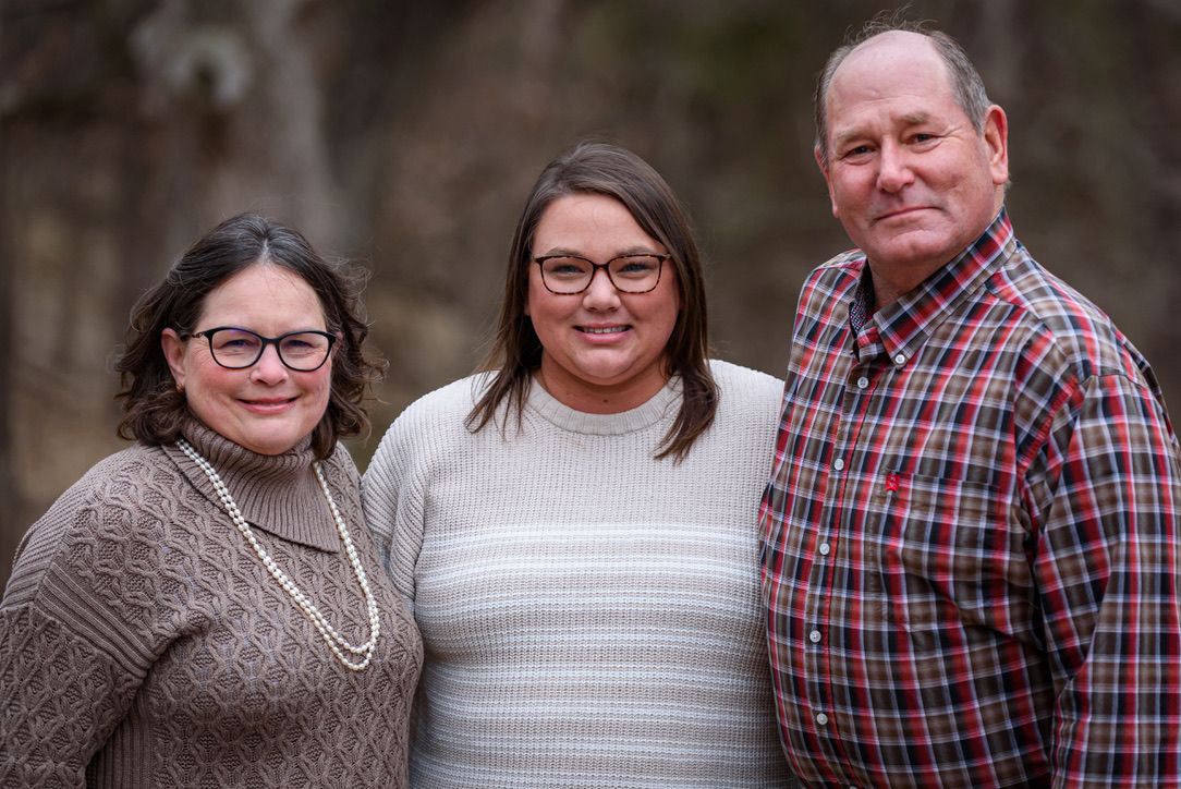 Three smiling adults pose together outdoors in casual attire for a family portrait photo.