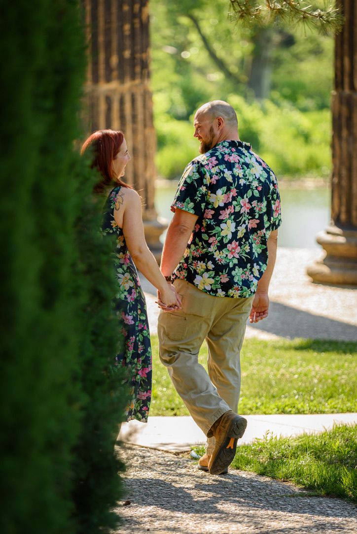 Couple holding hands walking through a lush green garden path, woman in floral dress and man in floral shirt.