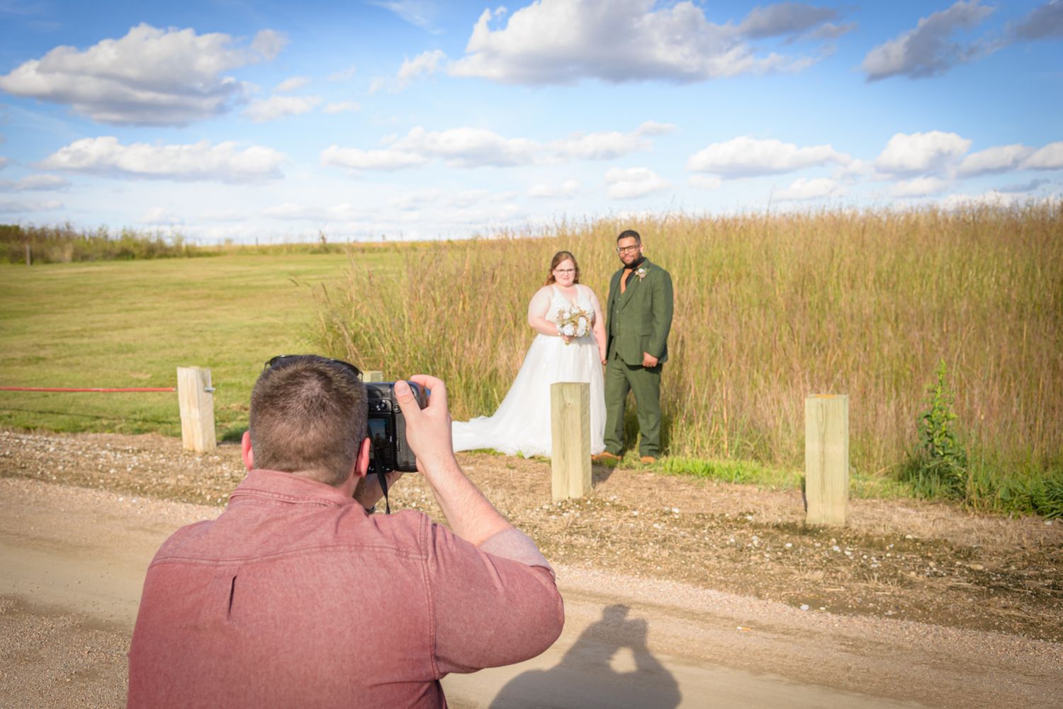 Photographer capturing wedding couple posing outdoors in a field with tall grass and blue sky. York, NE