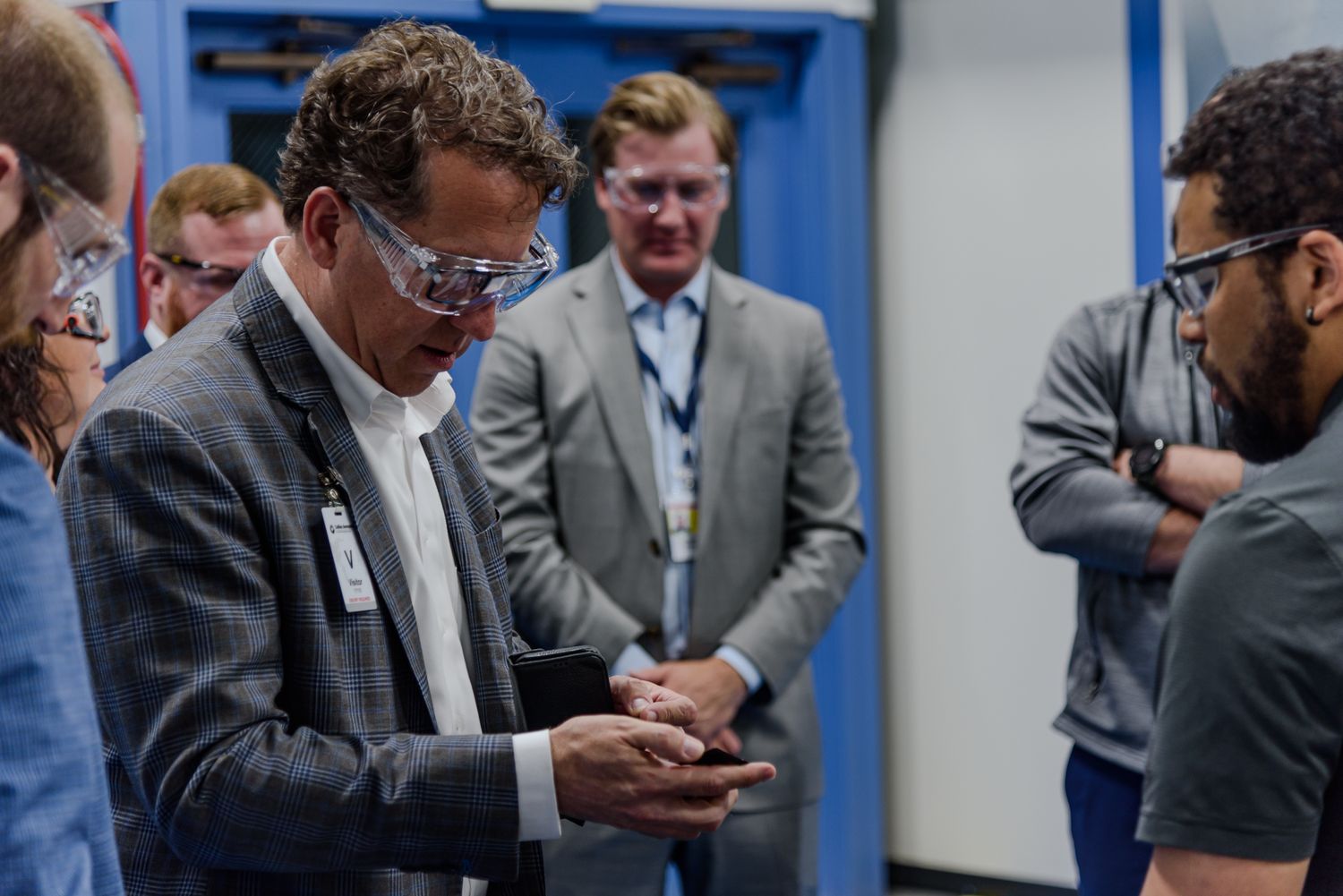 Professional wearing safety glasses examines object while colleagues observe in industrial facility.