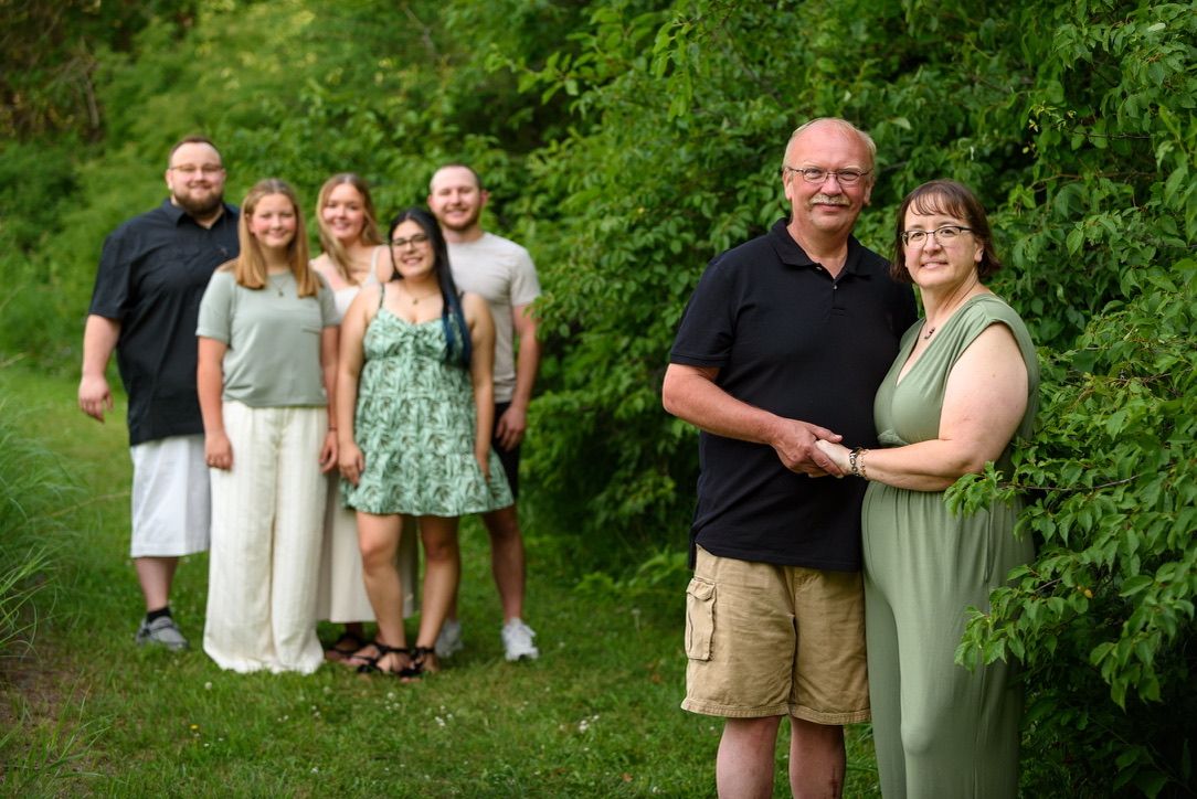 Family group photo outdoors with lush green trees, couple in foreground holding hands, young adults posing behind them.