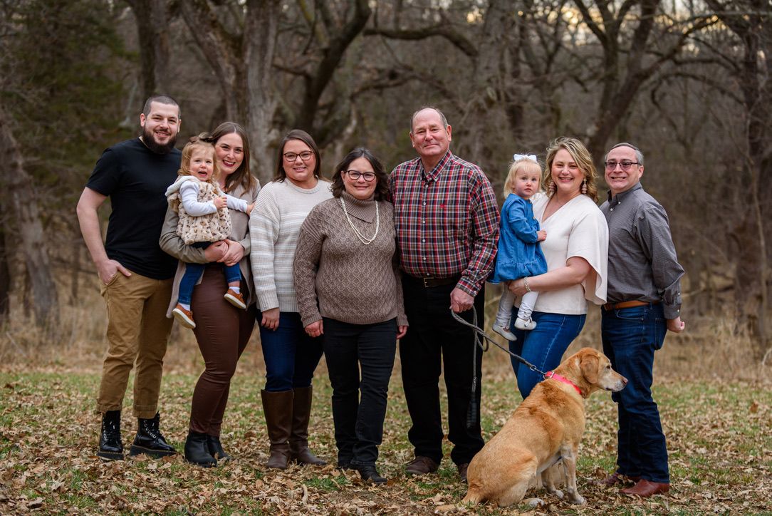 Family portrait with adults, toddler, and golden retriever dog posing outdoors in a wooded area during fall.