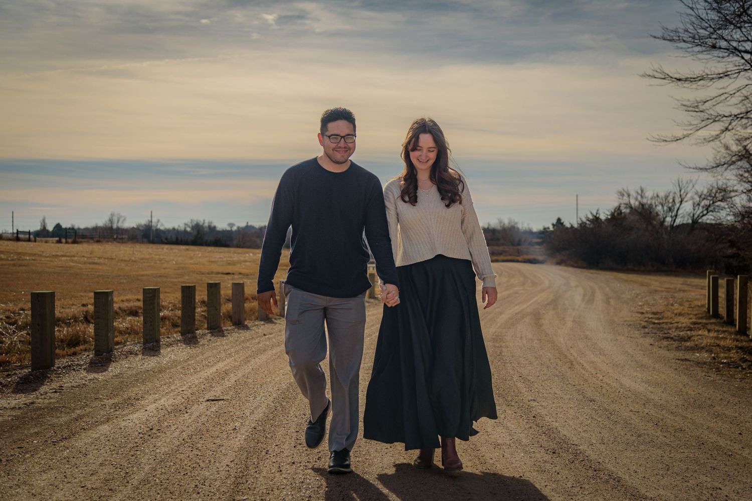 Couple walking hand in hand down rural dirt road at sunset with fence and fields. York, NE