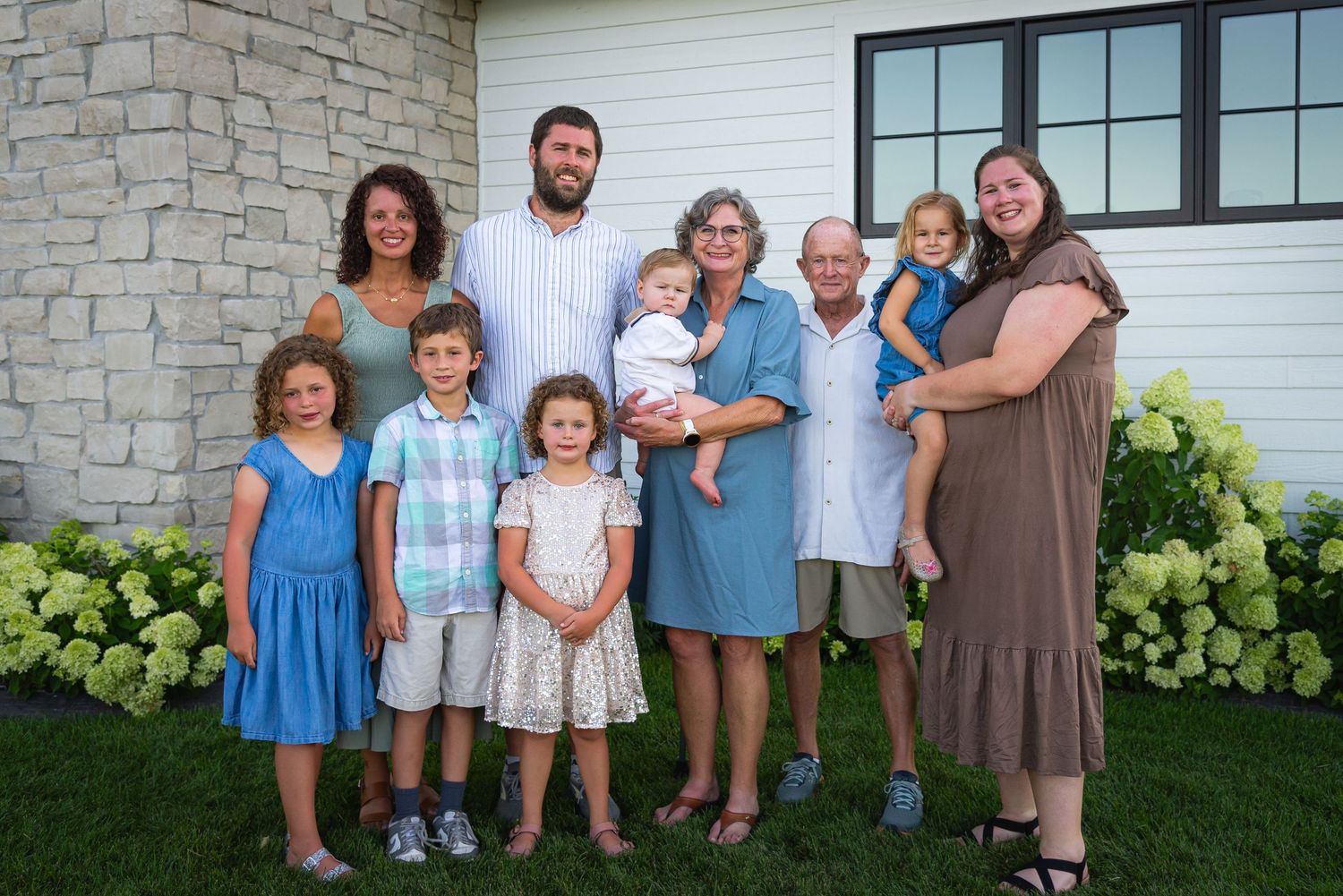Multi-generational family portrait outdoors, smiling adults and children posing together near a white building with green hydrangeas.