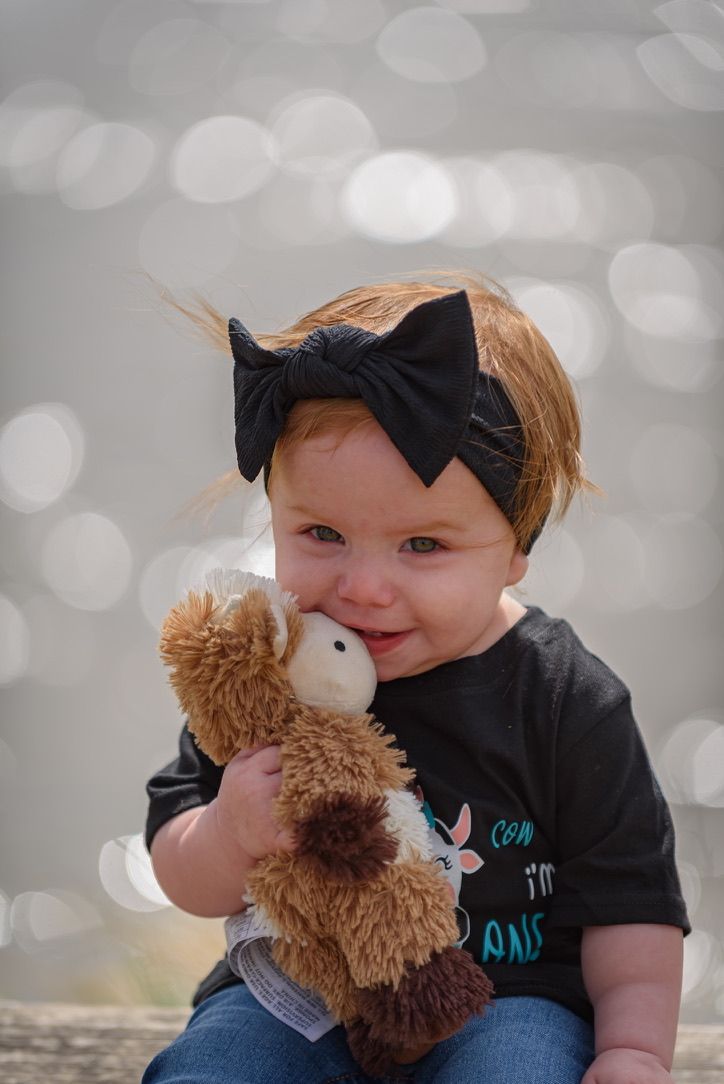 Smiling baby girl with black bow headband holding a brown teddy bear, wearing a black shirt, with bokeh background.