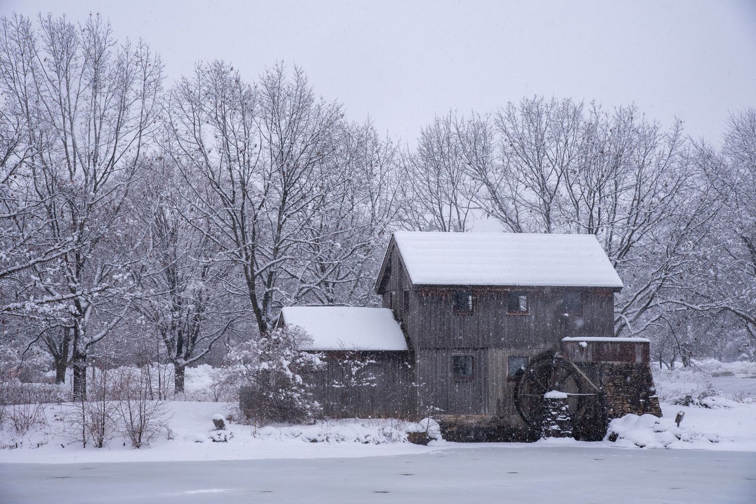 Snow-covered historic grist mill with water wheel beside frozen pond surrounded by frost-laden trees in winter landscape.