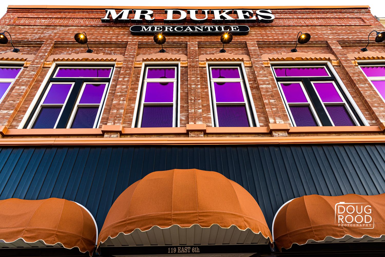 Mr. Dukes Mercantile brick storefront with purple-lit windows and orange awnings, photographed from below by Doug Rood.