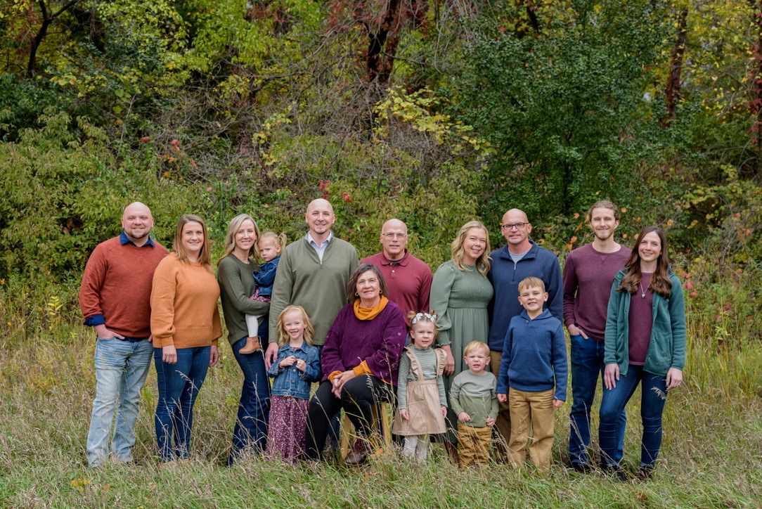 Large family group portrait outdoors in autumn, smiling adults and children posing in a green meadow with colorful fall foliage.