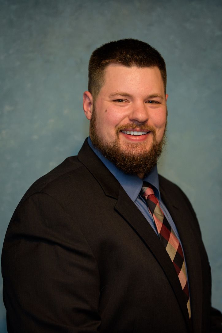 Professional man with beard smiling in dark suit and striped tie against blue background.