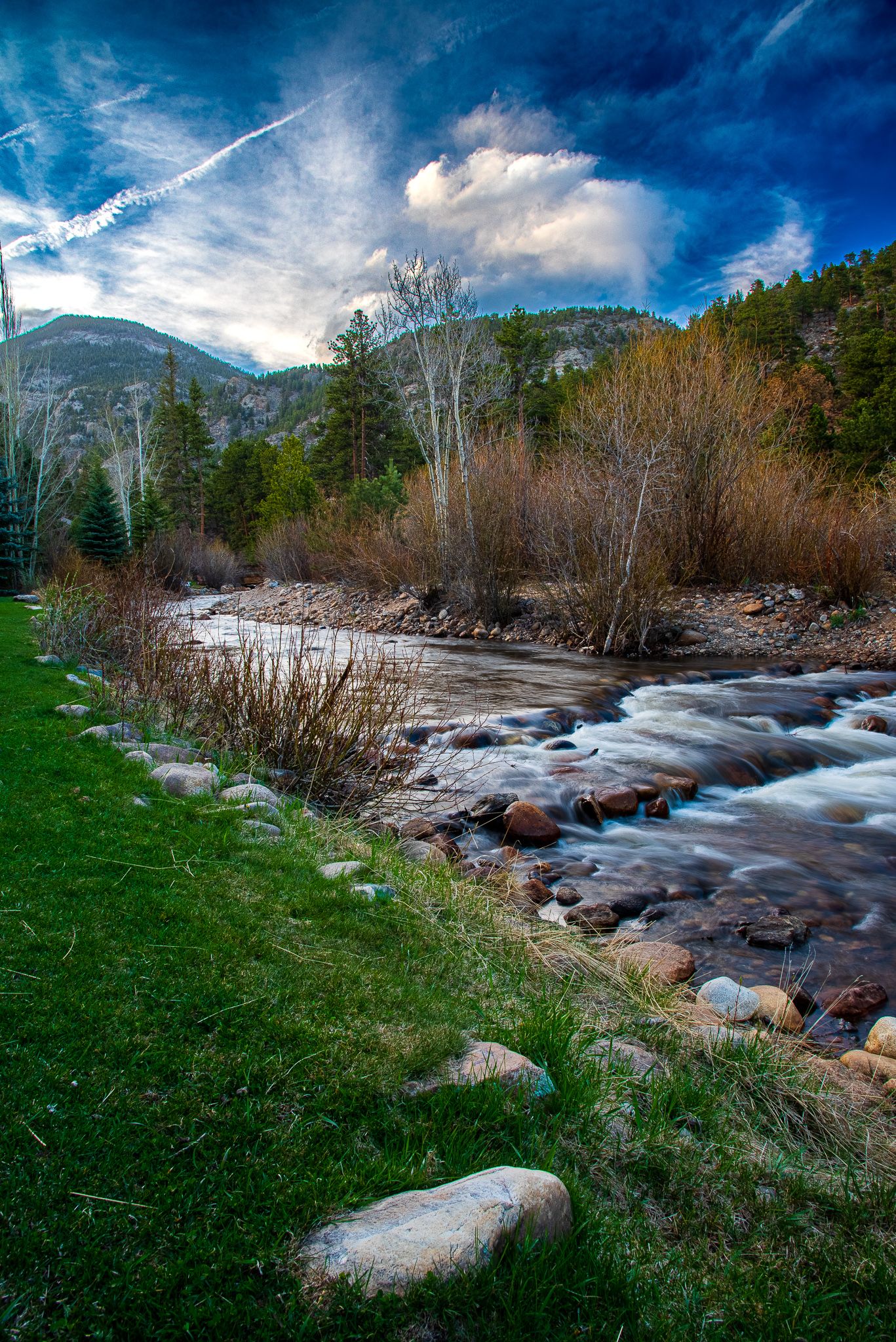 Rocky mountain stream flows swiftly past green grass banks with evergreen forest and dramatic cloudy sky backdrop.