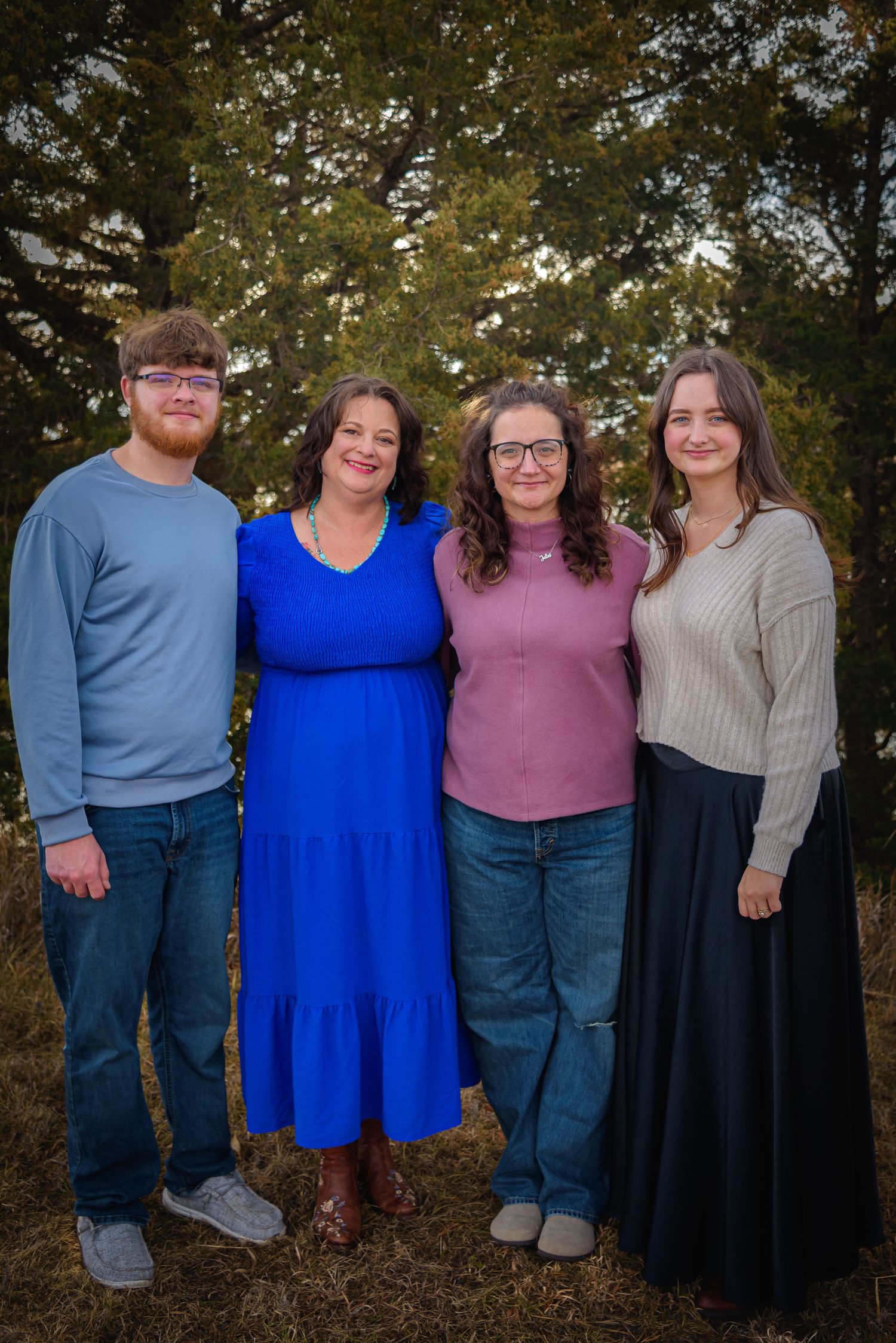 Family of four smiling outdoors among trees, wearing casual attire in blue, pink, and gray tones.