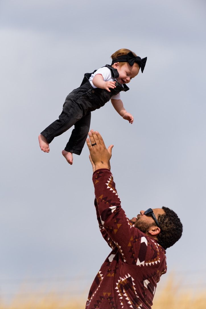 Father in red patterned shirt joyfully tosses smiling baby dressed in black outfit into the air outdoors.