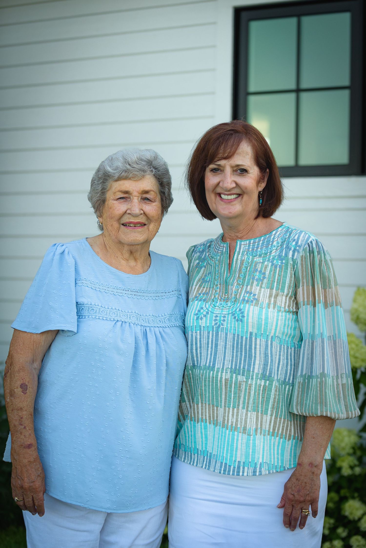 Two smiling women in blue tops pose together outdoors in front of a white house.