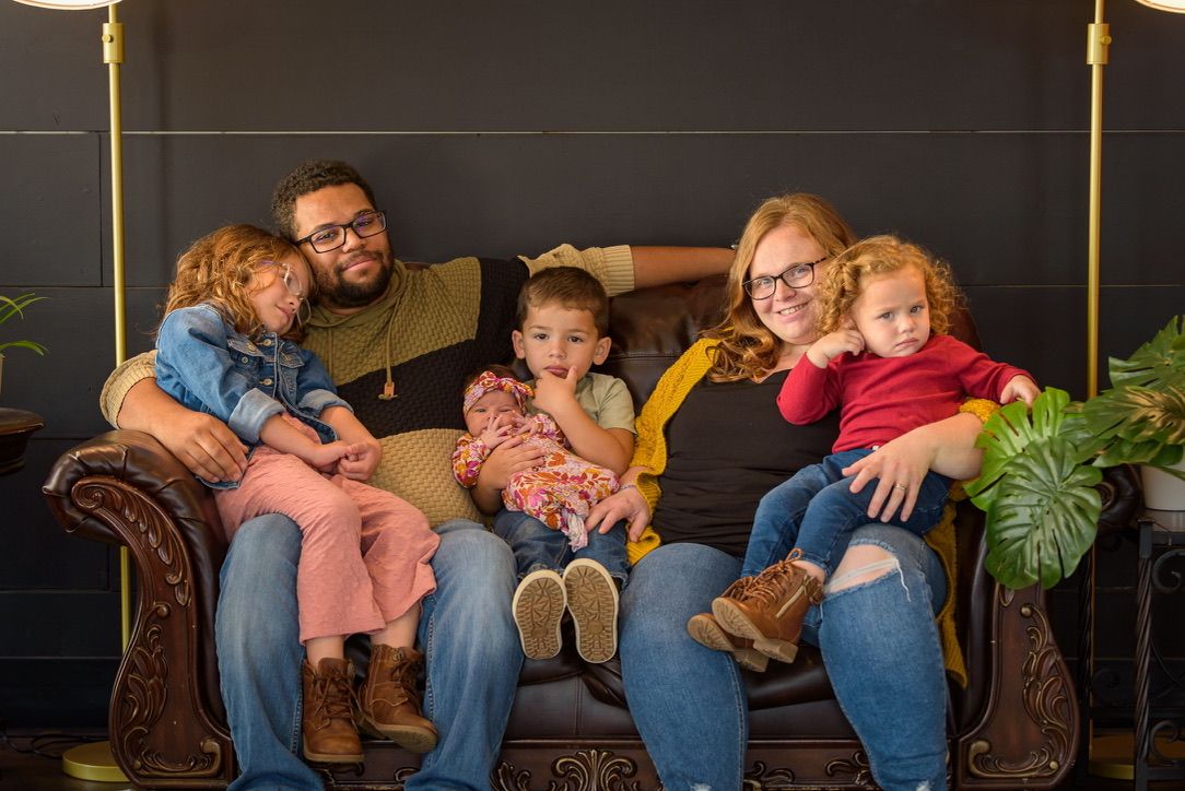 Happy family of five smiling together on a brown leather couch in a cozy living room setting.