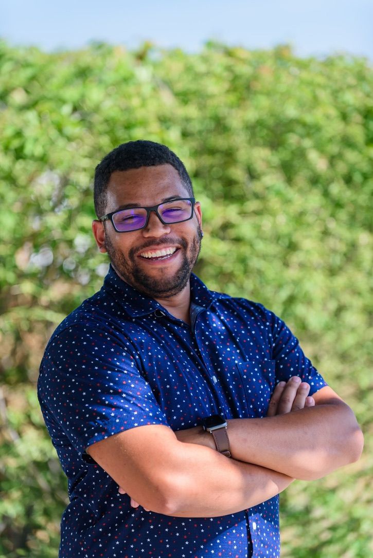 Smiling man with glasses and crossed arms wearing a navy blue polka dot shirt outdoors with green foliage background.