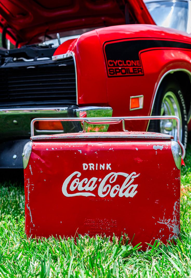 Vintage red Coca-Cola cooler on grass beside a classic red Cyclone Spoiler car at an outdoor auto show.