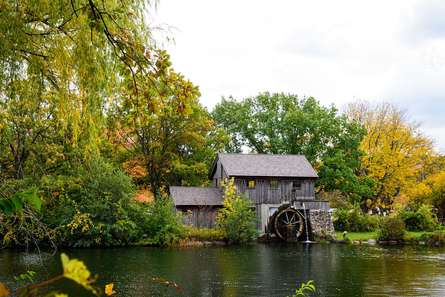 Historic wooden grist mill with water wheel beside a calm pond surrounded by colorful autumn trees.