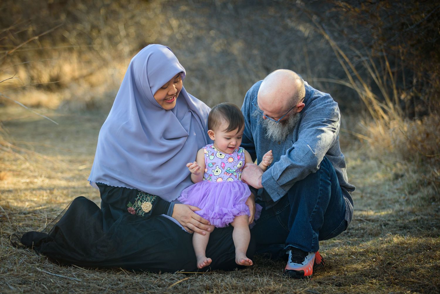 Grandparents kneeling outdoors with baby girl in purple tutu dress in natural setting. York, NE