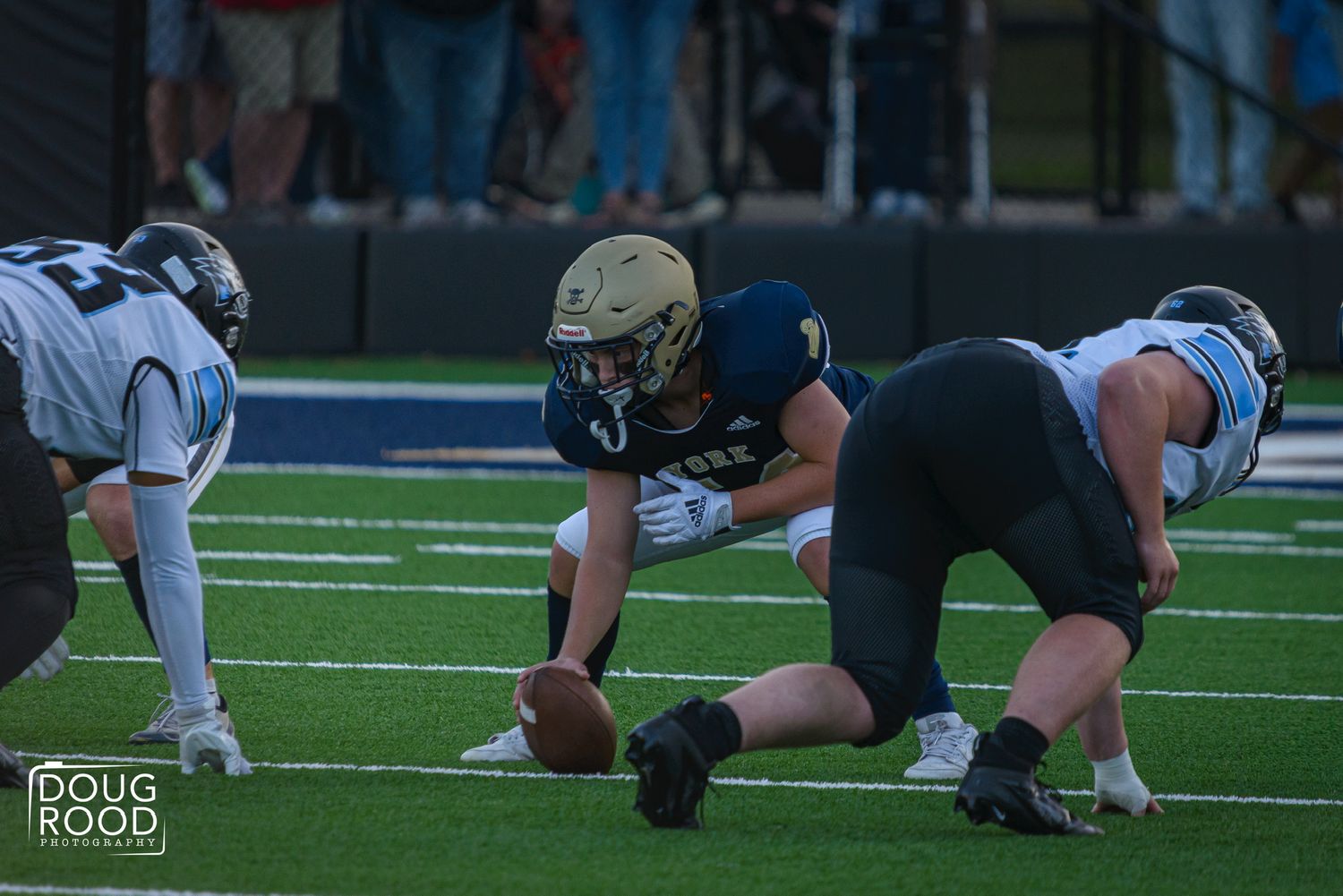 High school football player in gold helmet crouches over the ball at the line of scrimmage during a Friday night game.
