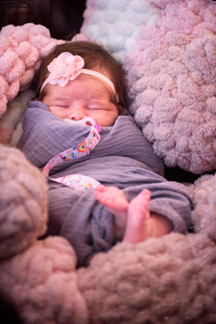 Sleeping newborn baby girl wrapped in gray swaddle with pink flower headband and pacifier surrounded by fluffy pink blankets.