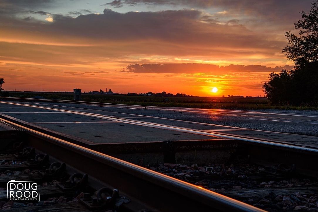 Railroad tracks leading toward a vibrant orange and red sunset over a flat horizon, photographed by Doug Rood.