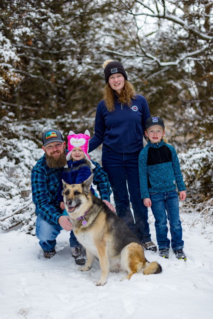 Family of four with German Shepherd dog posing in snowy winter forest, children holding pink heart balloon.