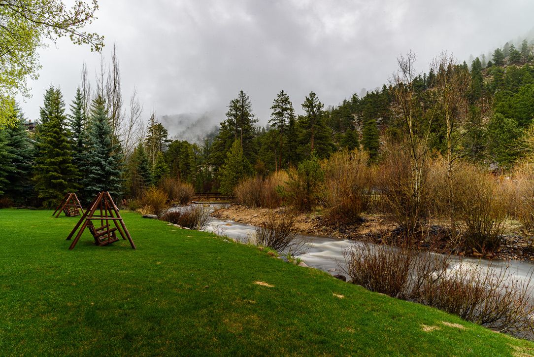 Lush green lawn with wooden playground equipment beside a flowing creek, surrounded by pine trees and misty mountains.