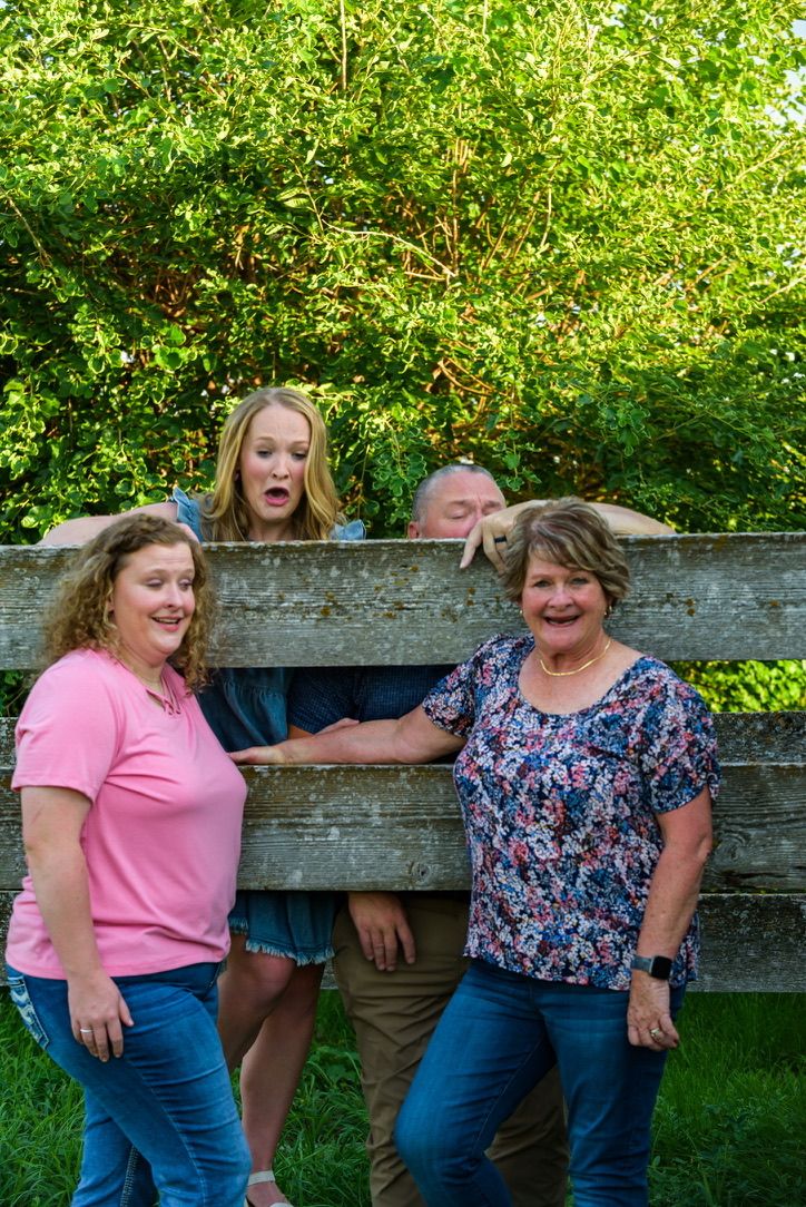 Family of four posing playfully behind a wooden fence outdoors, with lush green trees in the background.