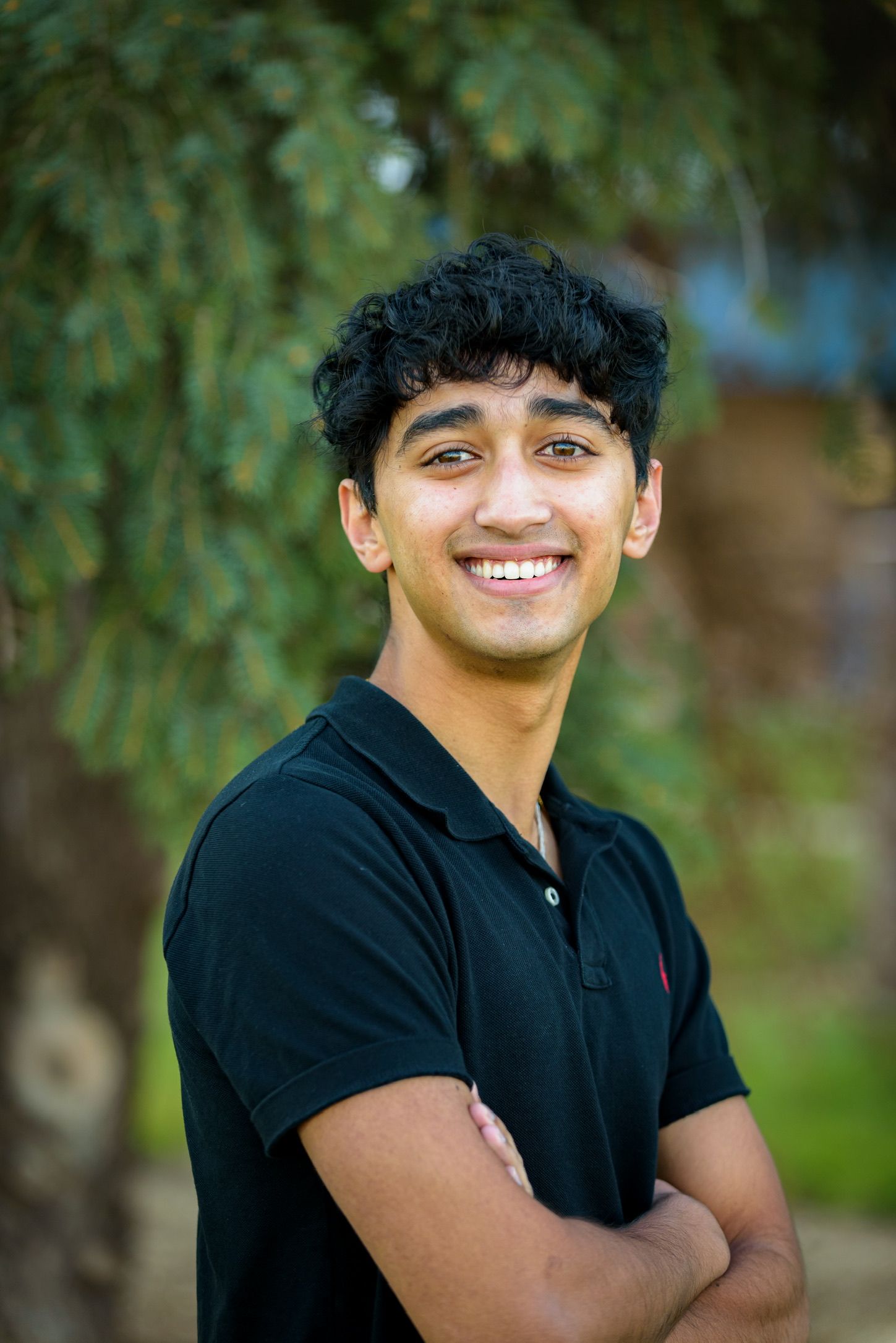 Smiling young man with curly black hair wearing a black polo shirt standing outdoors with arms crossed.
