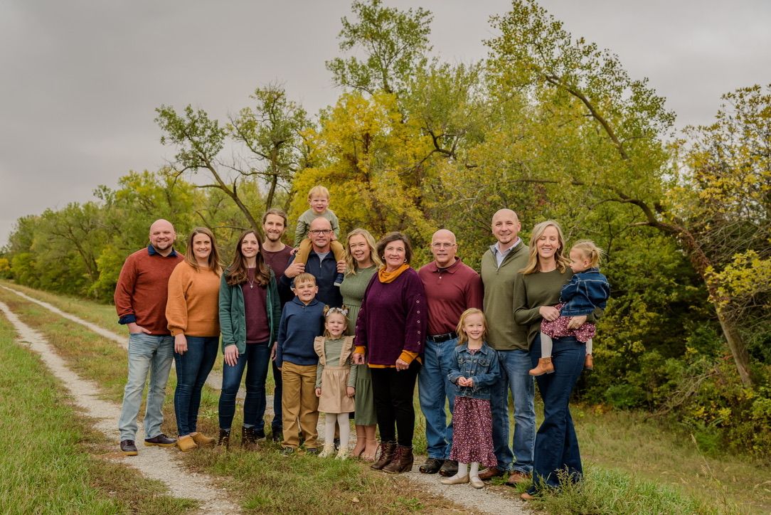 Large multigenerational family posing together outdoors on a gravel path surrounded by lush green and autumn trees.