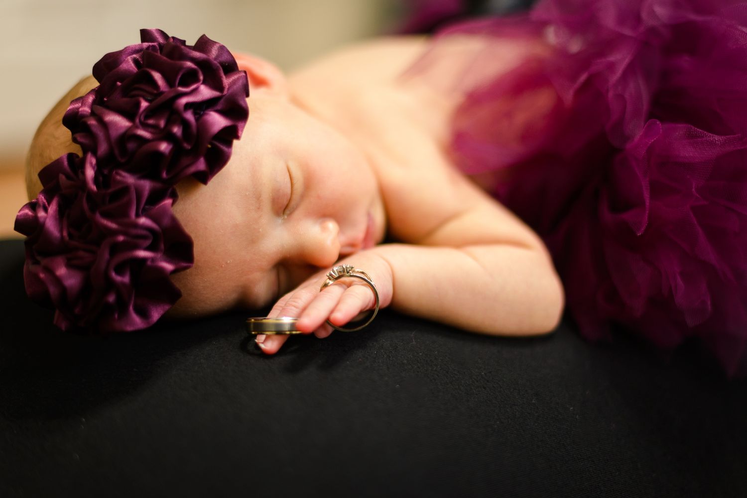 Sleeping newborn baby wearing a purple tutu and floral headband with a delicate ring on her finger.