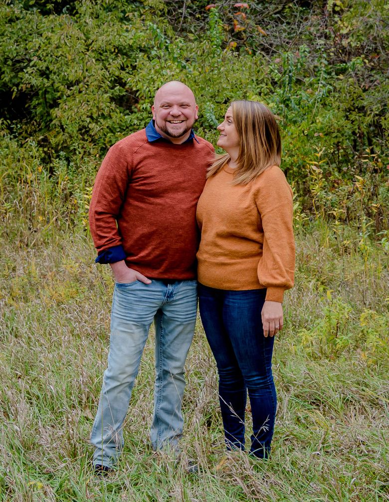 Smiling couple in fall sweaters standing in a grassy field with autumn foliage in the background.