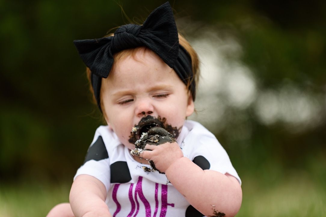 Adorable baby girl in cow print onesie and black bow headband eating cake with her hands outdoors.