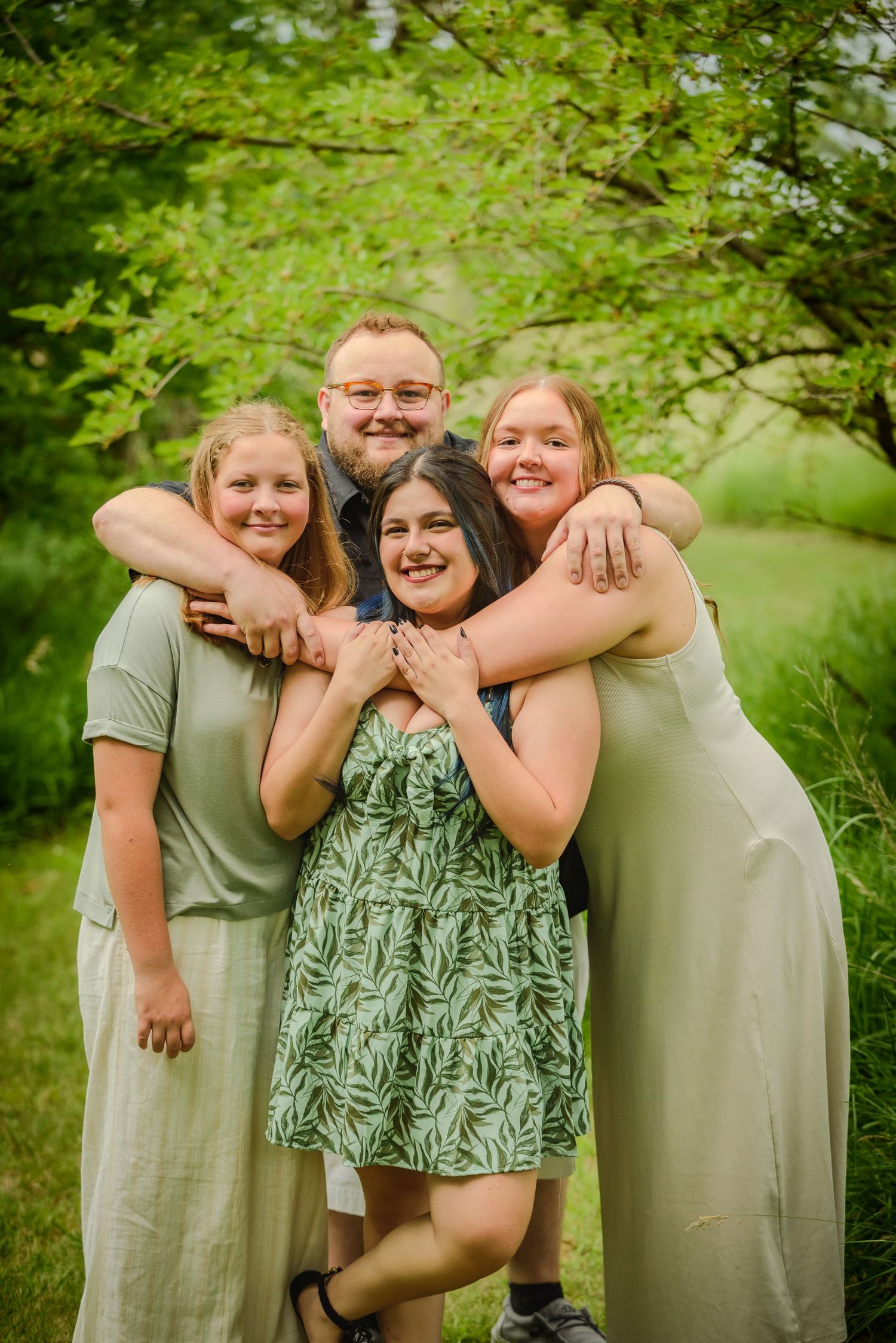 Family of four smiling and hugging outdoors in green nature setting, wearing coordinating sage green outfits.