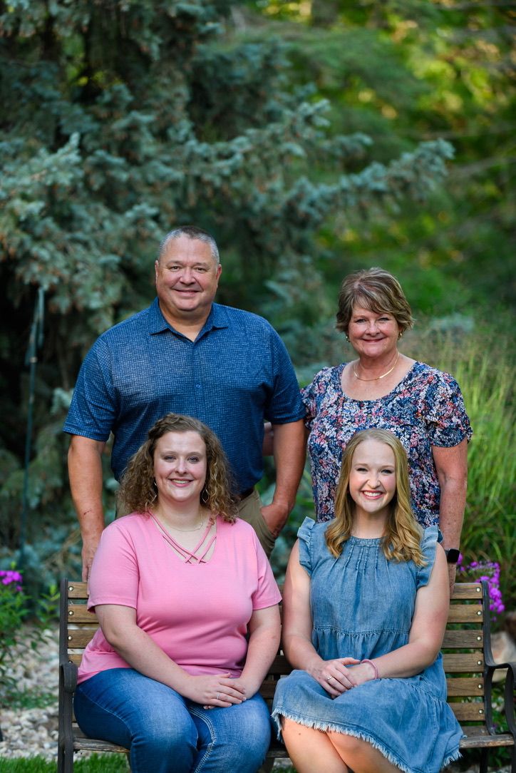 Family of four posing outdoors, two adults standing behind two young women seated on a bench, surrounded by lush greenery.
