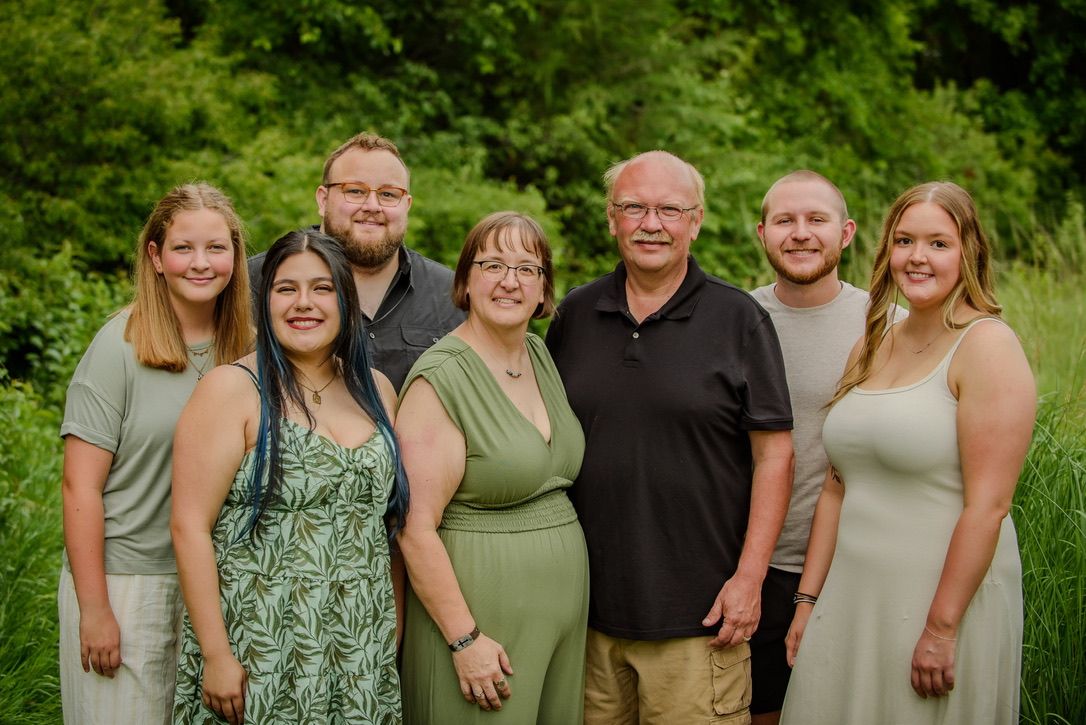 A joyful family of seven poses outdoors in front of lush green foliage for a professional portrait photo.